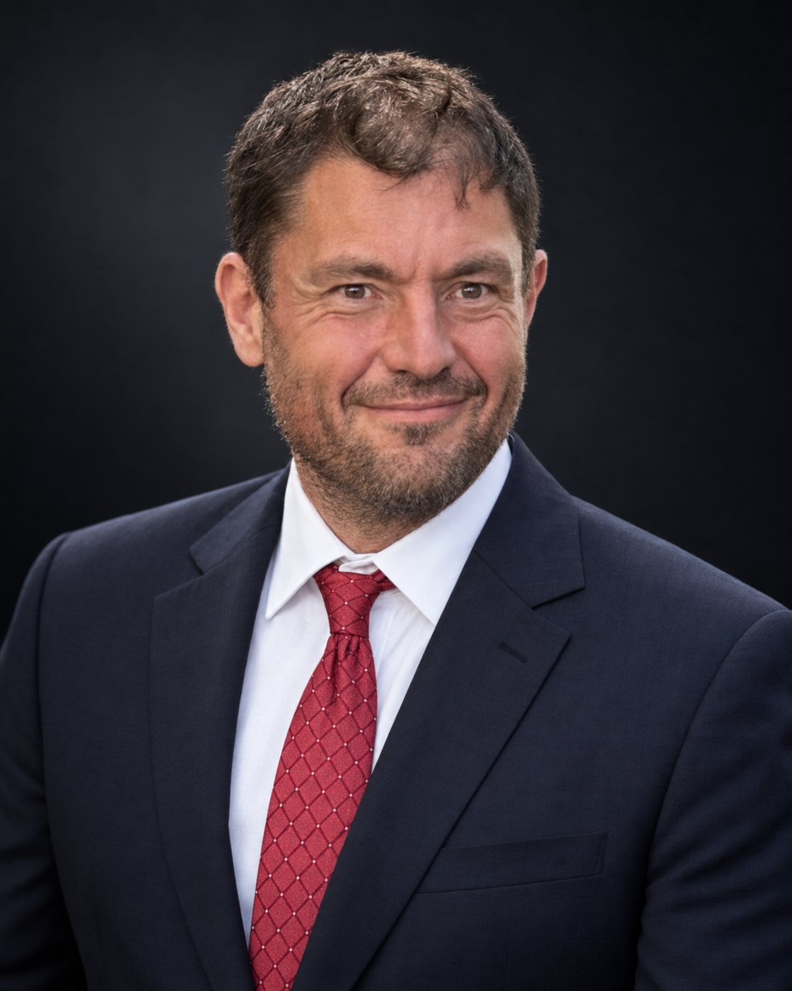Portrait of Nick Mermiges, Esq., in a navy suit, white shirt, and red tie